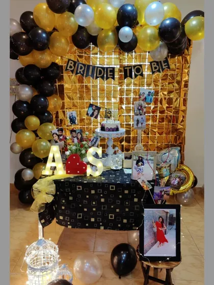 We help you celebrate the journey to the big day with stylish and personalized decorations. This 'Bride to Be' setup features a glamorous gold and black theme with a shimmer wall, balloon arch, and a dessert table personalized with photos.