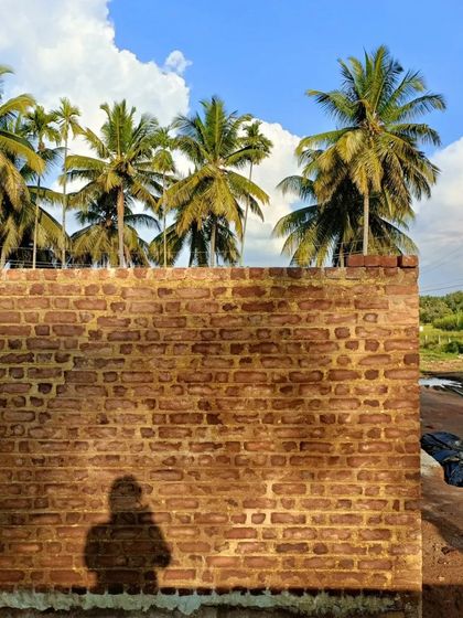 A newly constructed brick wall, built with a traditional lime mortar mix including jaggery and local herbs. The shadow of one of our team members falls on the wall, symbolizing our deep, personal connection to every project.