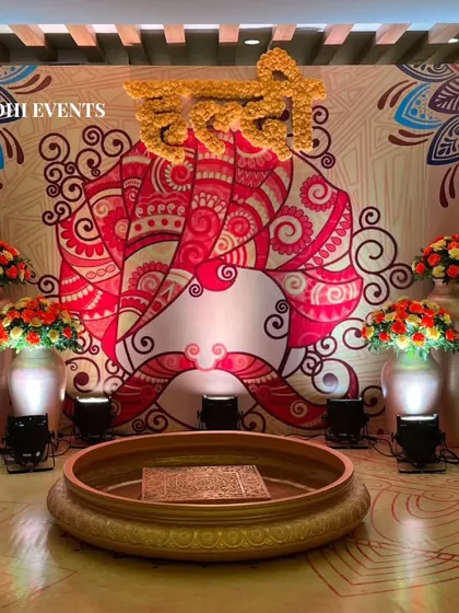 A traditional Haldi setup with a backdrop featuring classic Indian motifs and the word 'Haldi' written in flowers. The space is adorned with large vases of orange and white flowers.