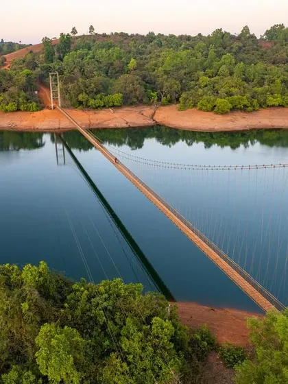 A hanging bridge stretches across a river in the Western Ghats. The reflection of the bridge in the calm water creates a beautiful symmetry.