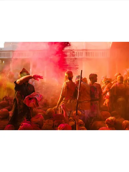 A crowd of people covered in colored powder during Holi, with one person throwing more color into the air. The hazy, colorful atmosphere conveys the immersive experience of the festival.