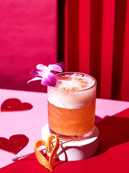 A frothy orange cocktail garnished with an orchid, styled with paper hearts against a bold red and pink striped background. This playful and romantic setup is ideal for a Valentine's Day menu.
