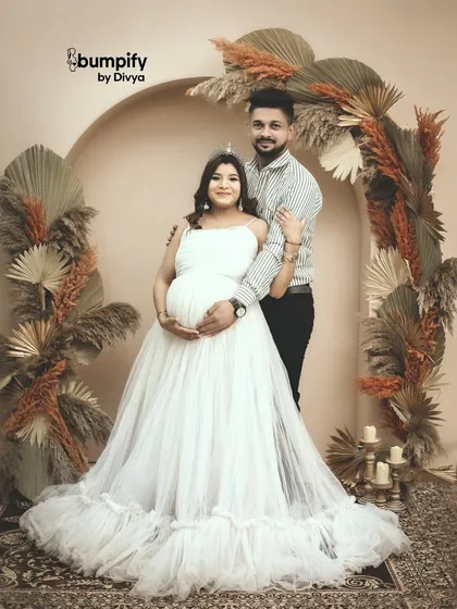 A royal boho look. The mom-to-be wears a tiara with her white gown, standing with her partner in front of a beautiful arch decorated with dried palm leaves and pampas grass.