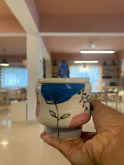 A lovely lidded jar, hand-painted with delicate flowers and topped with a little Buddha figure. This student's work is a beautiful blend of artistic skill and personal expression.