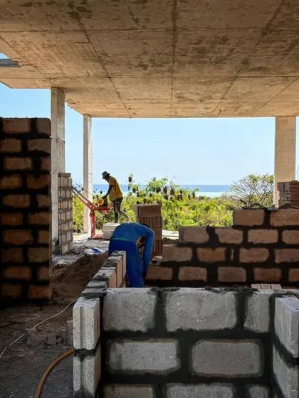 Artisans lay blocks for the walls of a new villa at Kizikula, with the ocean visible in the background. The open structure is designed for natural ventilation, essential for comfort in Zanzibar's tropical climate.
