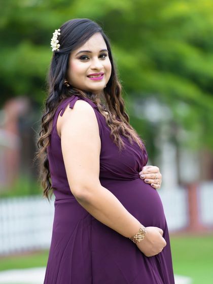 A close-up portrait in a park setting. The focus is on the mom-to-be's happy expression and the delicate floral hairpiece she wears.
