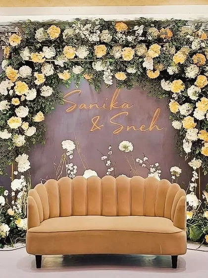 A dreamy engagement stage for Sanika and Sneh, featuring a beautiful arch of white and yellow roses against a soft grey backdrop. The personalized names and delicate floral details create a romantic and welcoming atmosphere.