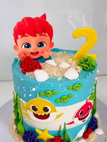 A close-up of the Baby Shark cake, focusing on the cute, red-headed figurine topper peeking out from the edible sand.
