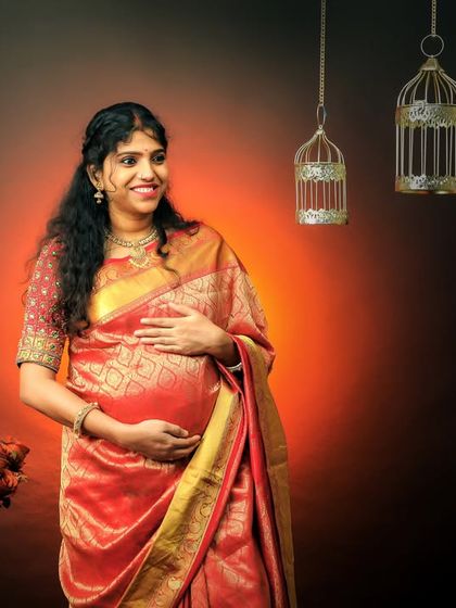 A smiling mother-to-be in a beautiful red and gold saree. The studio setup with hanging birdcages adds a touch of whimsical charm to this traditional portrait.