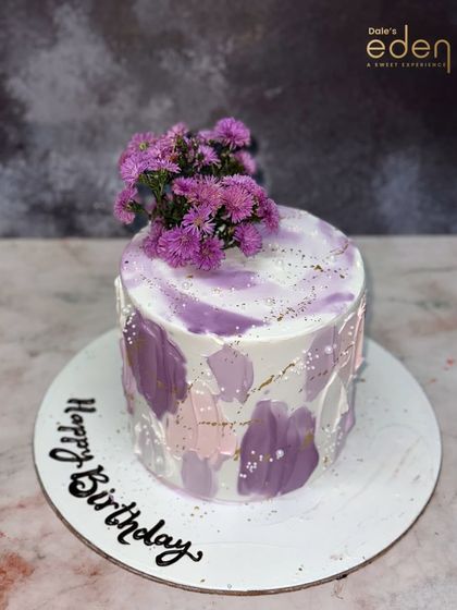A beautiful abstract design for a happy birthday. This cake features a watercolor-like effect with shades of purple and pink, topped with fresh purple flowers for a touch of natural beauty.