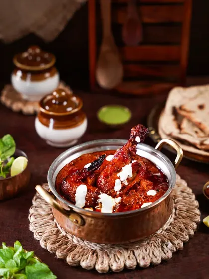 A delicious chicken curry in a copper pot, garnished with cream. The composition with traditional pickle jars and flatbread in the background tells a story of a complete, wholesome Indian meal.