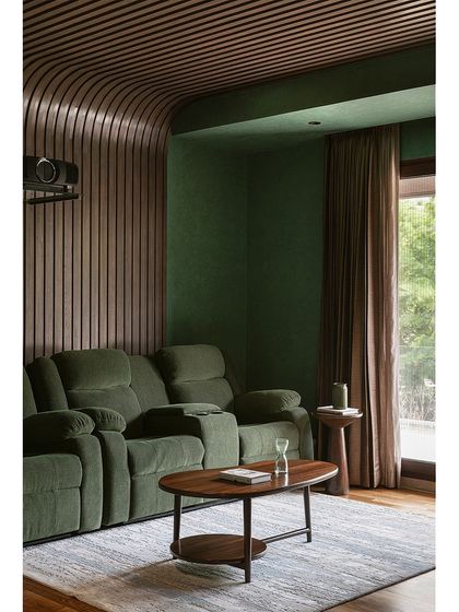 The media room at Tinted Tapestries, designed for ultimate comfort with plush green recliner sofas. The walls and ceiling are wrapped in curved, fluted wood panels for a cozy, cocoon-like effect and improved acoustics.