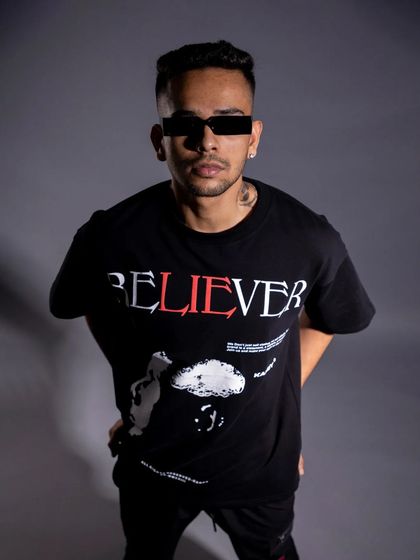 A classic studio portrait for a streetwear brand. The clean background and direct lighting put all the focus on the "Believer" graphic tee and the model's confident expression.