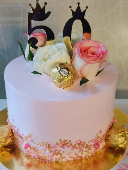 A chic and simple 50th birthday cake. The smooth pink frosting is accented with Ferrero Rocher chocolates, fresh roses, and a "50" topper with crowns.