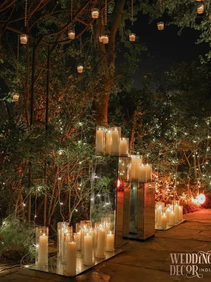 A romantic corner of an event, where mirrored pedestals covered in pillar candles create a beautiful reflection, amplifying the warm glow. Hanging candles from the trees above add to the enchantment.