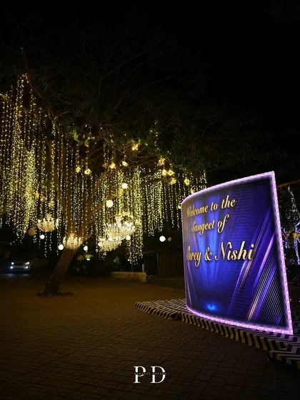 A modern, curved LED welcome sign for a 'Starry Night' Sangeet, set against trees adorned with hanging chandeliers and fairy lights.