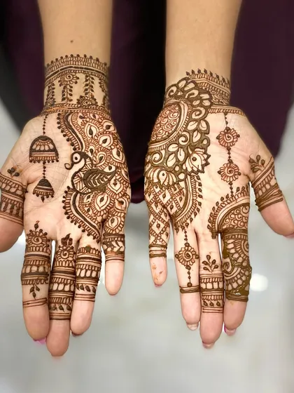 A stylish guest mehandi combining a peacock with jhumka motifs on one hand, and a floral mandala with hanging elements on the other.