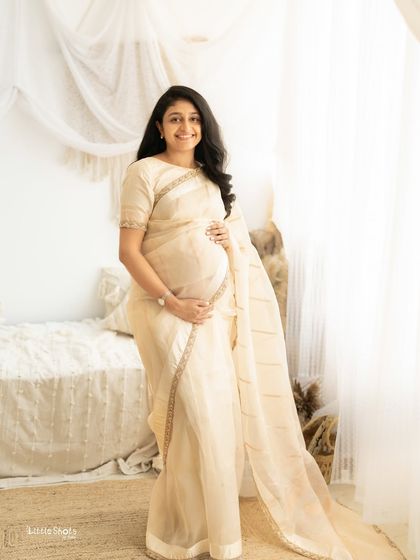 A full-length portrait of a smiling mother-to-be in a lovely cream saree. She stands in a bright and airy boho-themed room, her hand on her bump, looking happy and serene.