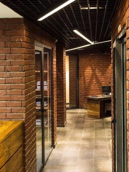 Inside the architect's office, the lighting design is functional and industrial. Linear fixtures are suspended from the open grid ceiling, providing clean lines of light that illuminate the workspace and complement the raw, honest materiality of the brick and wood.