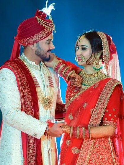 A picture-perfect couple. The groom is wearing a classic white sherwani with red accents, a red safa, and a beautiful gold necklace, coordinating perfectly with his bride.