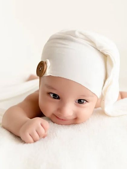 A smiling baby wearing a white sleepy cap lies on his tummy, looking at the camera.