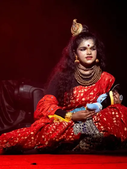 A tender moment portraying Devaki with the infant Krishna. The dramatic red lighting and the performer's intense expression convey the gravity and emotion of the scene.