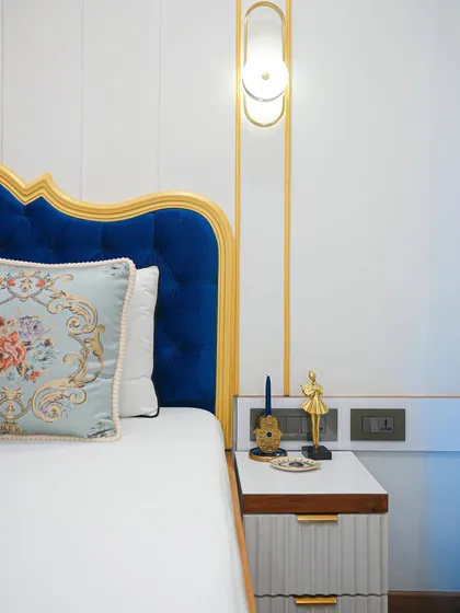A detail of the bedside, where a modern gold wall sconce is juxtaposed with the classic, ornate headboard and embroidered cushion.