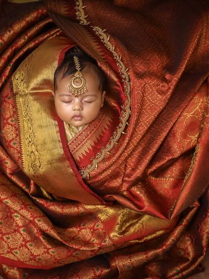 This stunning portrait captures a newborn wrapped in the rich fabric of a traditional red and gold saree. The intricate details of the fabric create a luxurious and culturally rich setting for this peaceful moment.