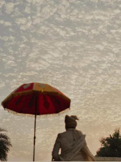 An artistic shot of the groom under a traditional umbrella against a dramatic, cloud-filled sky. This image captures a sense of grandeur and cultural richness.