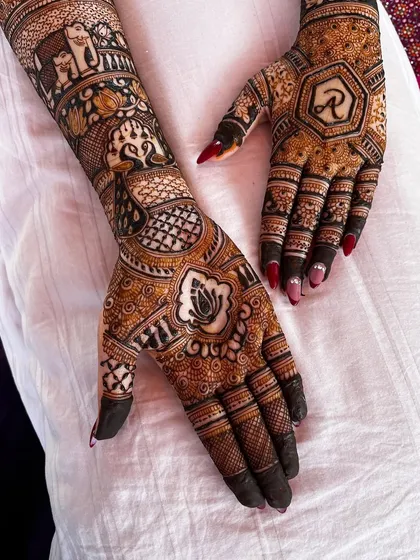 A close-up of a bridal mehendi featuring a lotus motif and the groom's initial 'R' inside a hexagon. The surrounding design is filled with intricate details and shading.