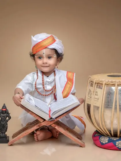 A sweet portrait of a baby boy dressed as a Varkari, sitting with a holy book and a tabla. The soft, neutral background keeps the focus on the intricate details of the traditional setup.