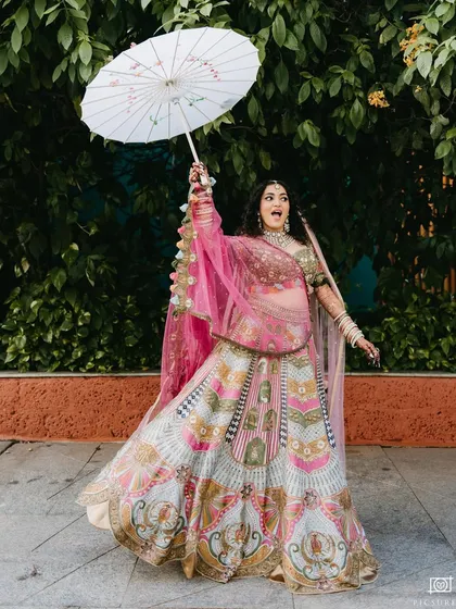 A joyful bride in a colorful pink lehenga twirls with a white parasol, her expression full of happiness and excitement.