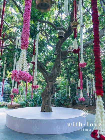 A 'Katté' or traditional seating platform built around a tree, decorated with hanging floral malas and temple bells for guests to relax under.