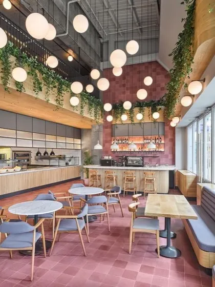 We designed this spacious, double-height cafe to feel vibrant and full of life. The design features a bold pink tiled floor, an array of spherical pendant lights for dramatic effect, and hanging greenery to soften the industrial architecture, creating an energetic and welcoming atmosphere for customers.