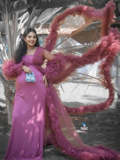 A stylish outdoor portrait featuring a dramatic ruffled gown. The mom-to-be holds a pair of baby shoes, blending high fashion with a sweet, personal touch.