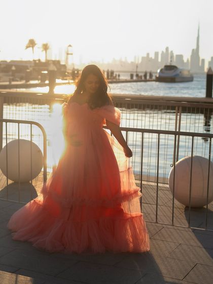 A dreamy, sun-flared shot with the Dubai skyline in the distance. The mom-to-be looks ethereal in her pink gown, walking along the waterfront as the sun sets.
