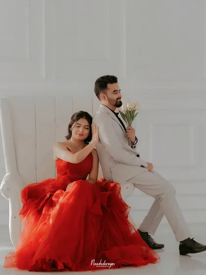 A posed, elegant shot of a couple sitting on a large white chair, the bride in a flowing red dress.