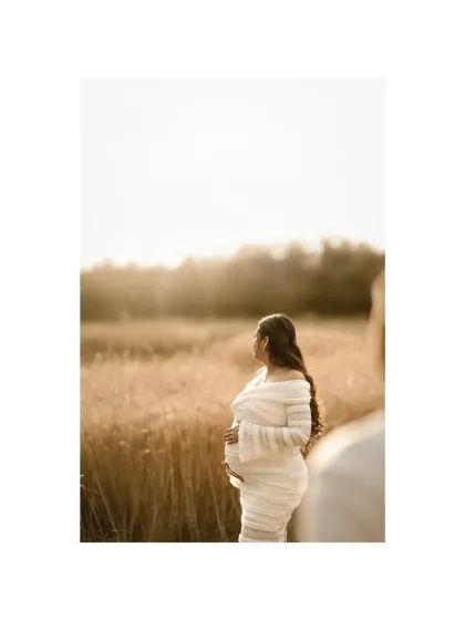A beautiful over-the-shoulder shot, with the partner blurred in the foreground, creating depth and a sense of shared perspective. The mother-to-be looks out over the golden field, lost in thought.