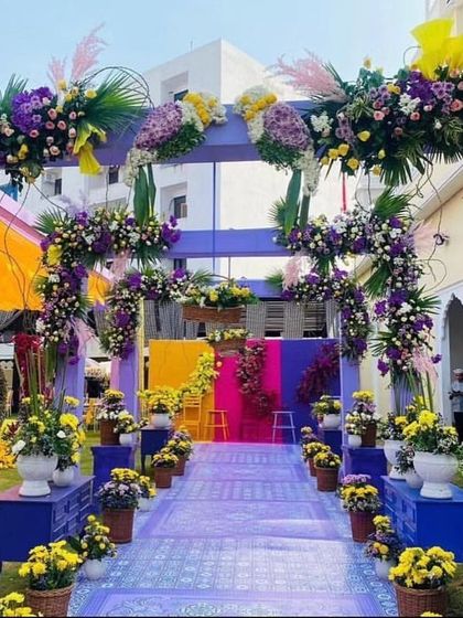 A grand floral walkway designed for a Haldi ceremony. The path is lined with potted flowers and leads through a series of arches adorned with purple and yellow floral arrangements.
