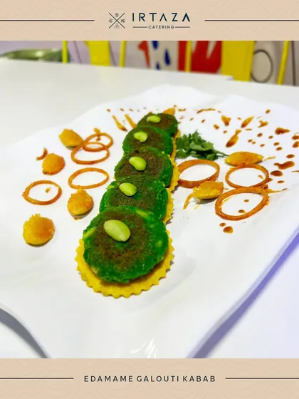 An artfully plated Edamame Galouti Kebab. The vibrant green of the kebab is contrasted with swirls of sauce and crispy onion rings, showcasing a dish that is as much a work of art as it is delicious.