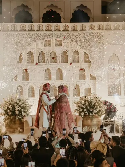 A varmala ceremony against a backdrop of a palace-like structure. The cold pyros and the crowd of cameras make this feel like a celebrity wedding.