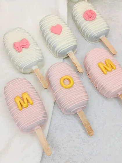 Our "MOM" cakesicles, decorated in pink and white chocolate, are a cute and delicious part of our Mother's Day hamper.