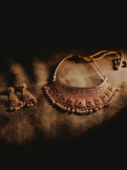 A close-up, artistic shot of the bride's wedding jewelry. The dramatic lighting highlights the intricate design of the gold necklace and earrings, essential elements of the bridal look.