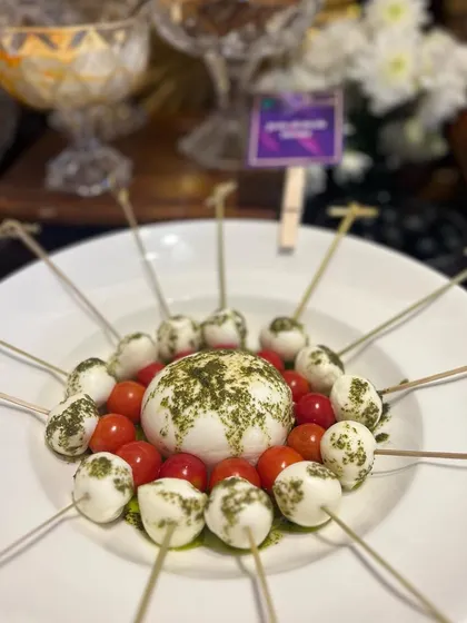 A modern presentation of a Caprese salad. Cherry tomatoes and mozzarella balls are skewered and arranged around a ball of fresh burrata, drizzled with pesto.