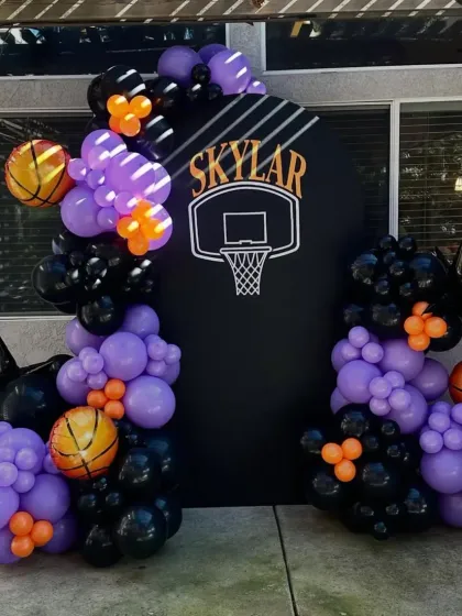 A dynamic basketball-themed party with a custom backdrop featuring a hoop, and a balloon arrangement in purple, black, and orange.