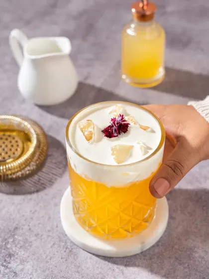 A hand holds a beautiful orange cocktail with a creamy white foam top, garnished with a delicate flower. The bright, clean lighting and simple props create a fresh and elegant feel for this drink photography.