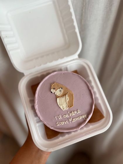 A sweet bento cake for a mom-to-be. This design features a hand-drawn bear with the message "I'll be here soon Mommy" on a lavender-colored buttercream base.