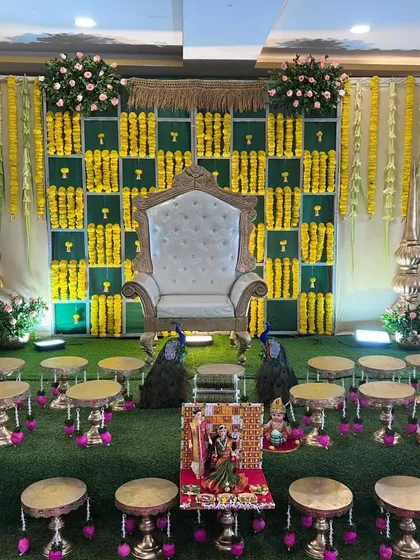An elaborate and traditional baby shower stage, rich in cultural details. The decor includes a checkered backdrop of green panels and yellow marigolds, a regal white throne, hanging floral garlands, and brass accents, all set on a green turf for an authentic feel.