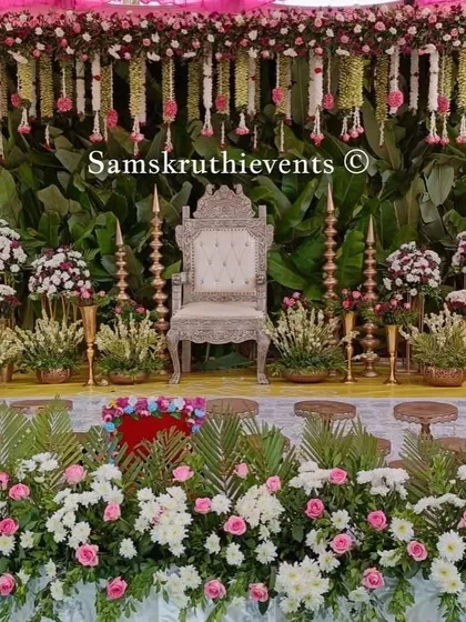 A grand stage setup with a banana leaf backdrop. The decor is enhanced with an ornate silver throne, traditional brass lamps, and a lush border of white and pink flowers.