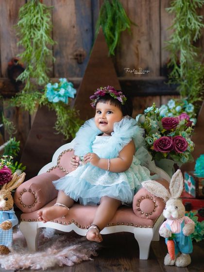 A sweet baby girl in a fluffy blue dress, sitting on a miniature pink sofa. The woodland theme with bunny props adds a touch of fairytale magic.